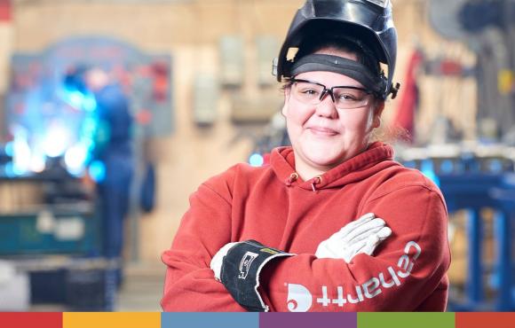 A young First Nations woman is standing with her arms crossed, smiling while standing in the welding bay. She is wearing a welding shield on top of her head and there is a blurred out person welding with sparks flying in the background. 