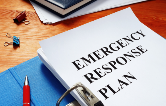 A three ring binder lays open on a wooden table with a red pen and some paper clips strewn about. The title on the open page reads "Emergency Response Plan" 